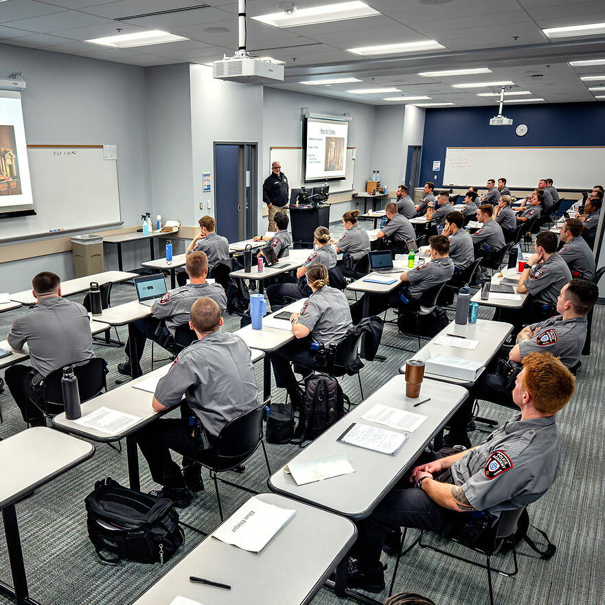 A group of uniformed individuals sits at desks in a classroom, attentively listening to a presenter at the front.